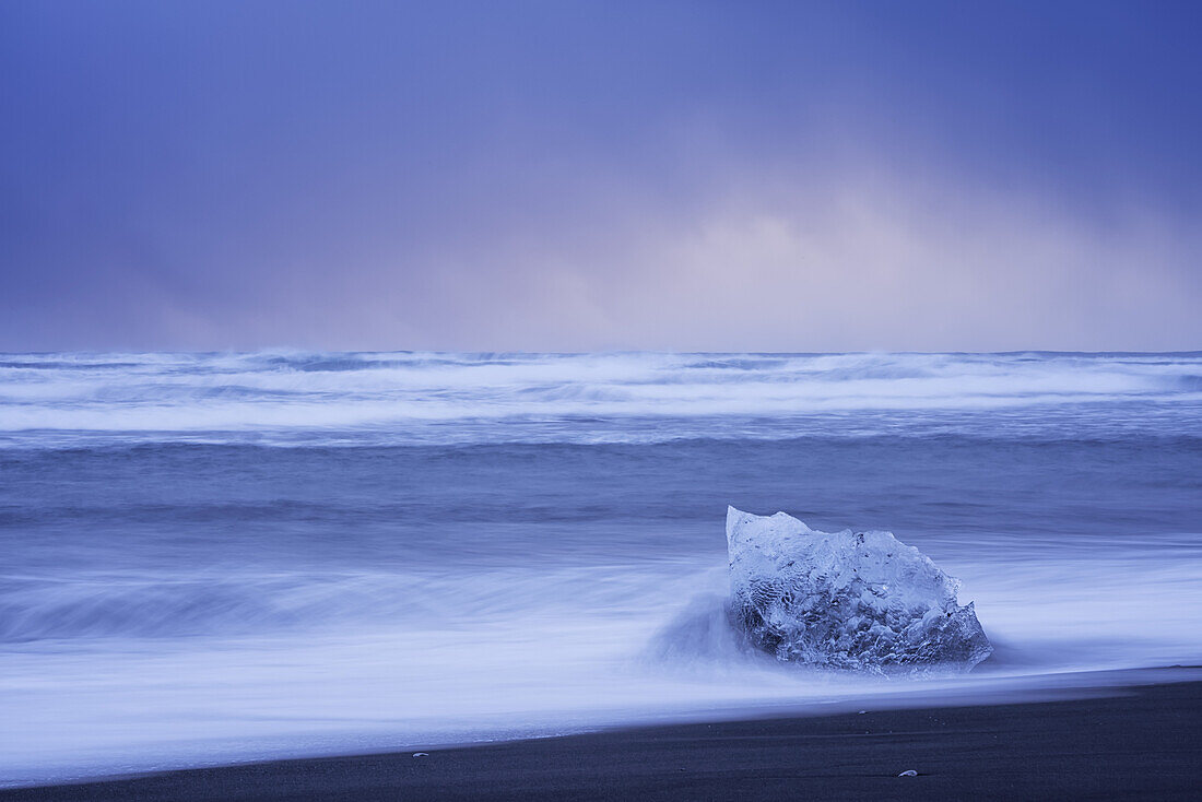 Ein Stück Eis, das von den Wellen an der Südküste Islands getroffen wird, mit stürmischem Himmel im Hintergrund; Island