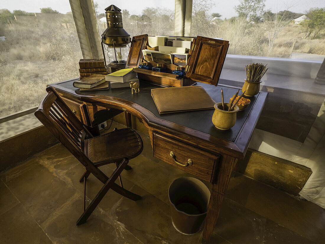 Schreibtisch im Foyer eines Zeltes, umgeben von Fenstern mit Blick auf eine Landschaft aus trockenen Büschen und Bäumen; Rajasthan, Indien.