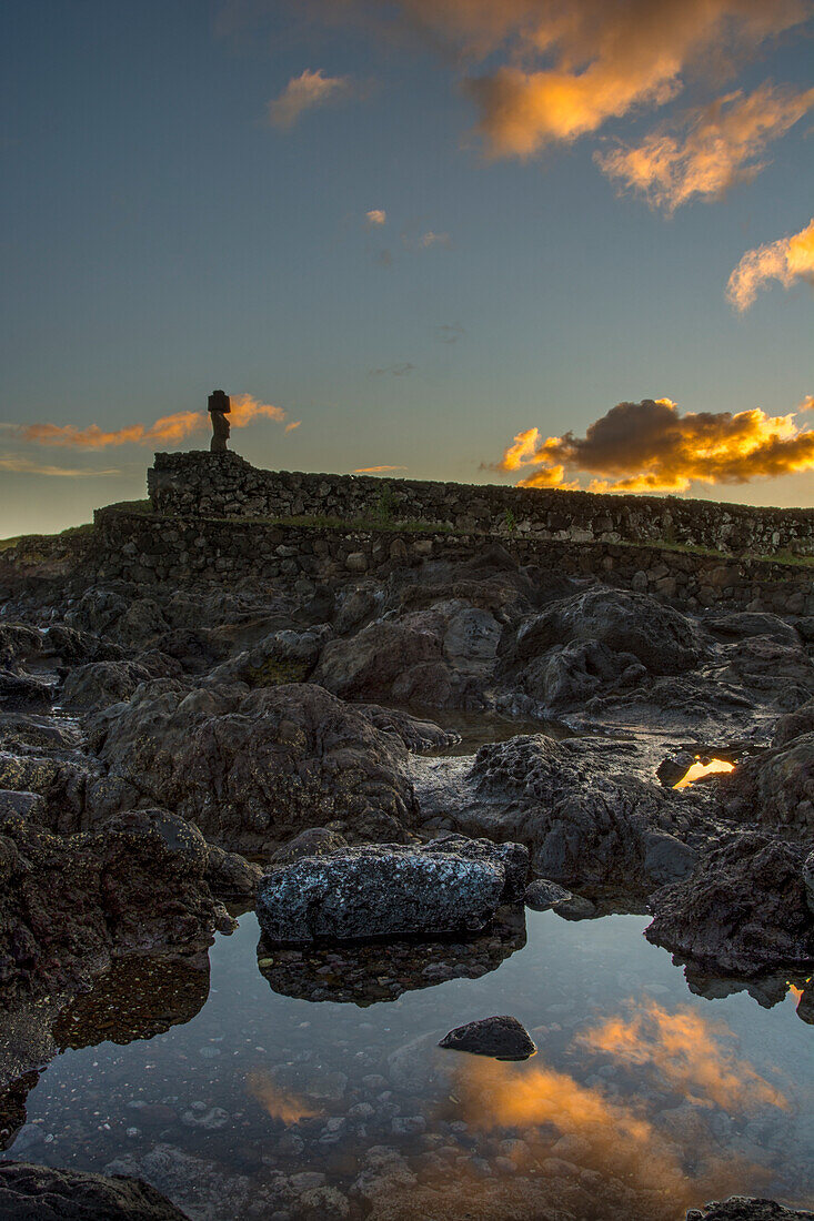 Ein einzelner Moai in der Ferne vor dem von der aufgehenden Sonne beleuchteten Himmel, der sich in einem Wasserbecken im Vordergrund spiegelt; Osterinsel, Chile