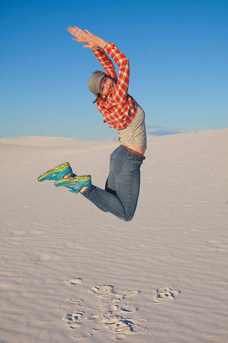 A young woman leaps in the air on the white sand with blue sky, White Sands National Monument; Alamogordo, New Mexico, United States of America