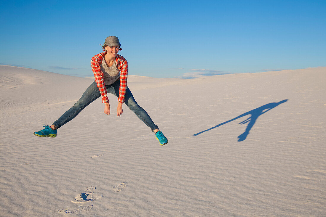 Eine junge Frau springt in die Luft auf dem weißen Sand mit blauem Himmel, White Sands National Monument; Alamogordo, New Mexico, Vereinigte Staaten von Amerika