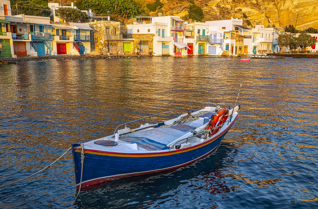 Ein im Vordergrund vertäutes Boot mit bunt bemalten Geländern und Häusereingängen entlang der Uferpromenade bei Sonnenuntergang; Milos, Griechenland