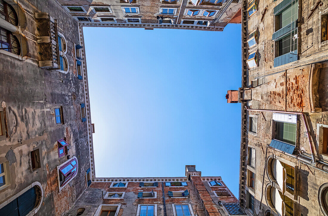 View of the blue sky above from directly below with the weathered walls of residential buildings on all four sides; Venice, Italy
