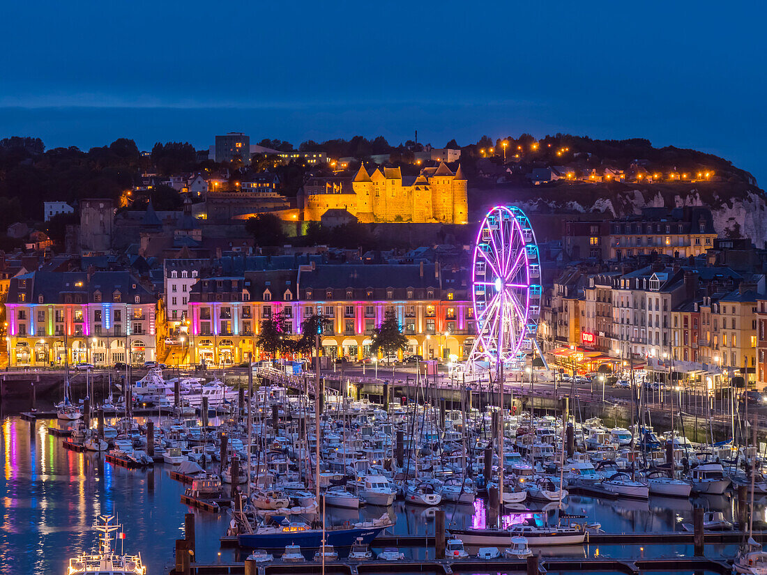 Harbour full of boats and an illuminated ferris wheel at night with lights illuminating the Chateau de Dieppe castle on the hillside; Dieppe, Normandy, France