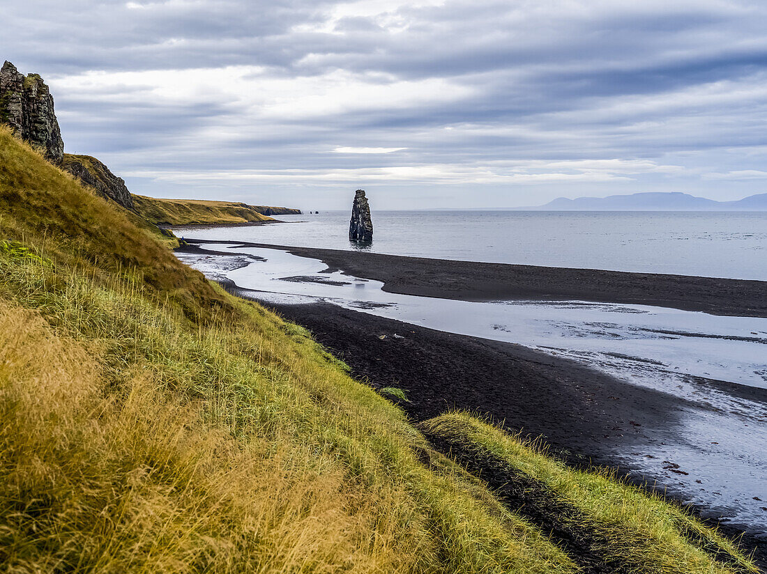 Tall rock formation and grassy slopes along the shoreline of a fjord; Iceland