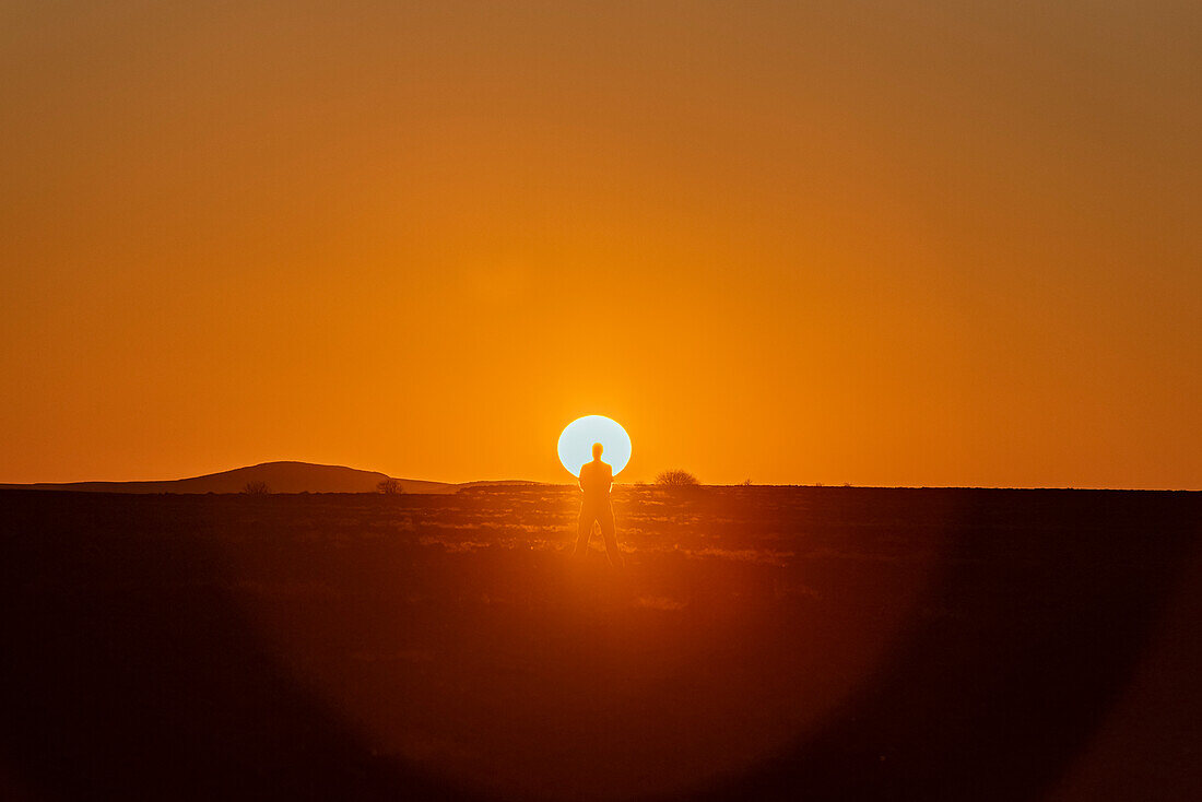 Mann steht im Gegenlicht der hellen, glühenden Sonne, die bei Sonnenuntergang hinter dem Horizont versinkt, in Aluvlei, Namib-Naukluft-Nationalpark; Namibia.