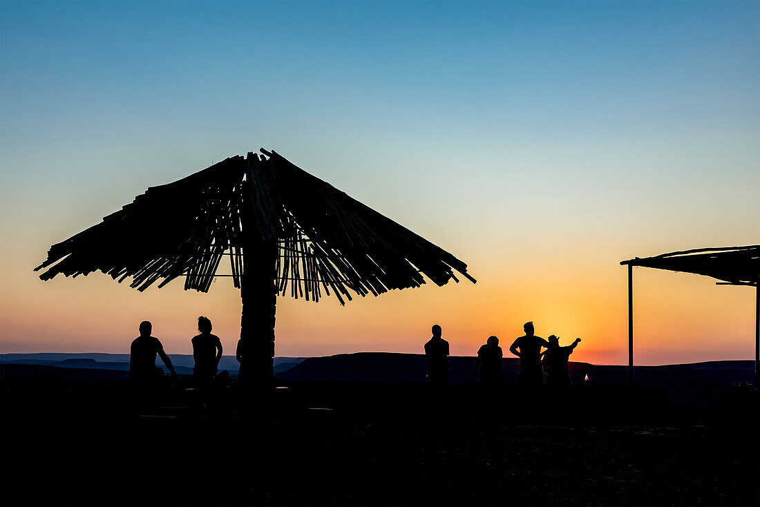 Silhouetted tourists and umbrella at sunset, looking out over Fish River Canyon; Namibia