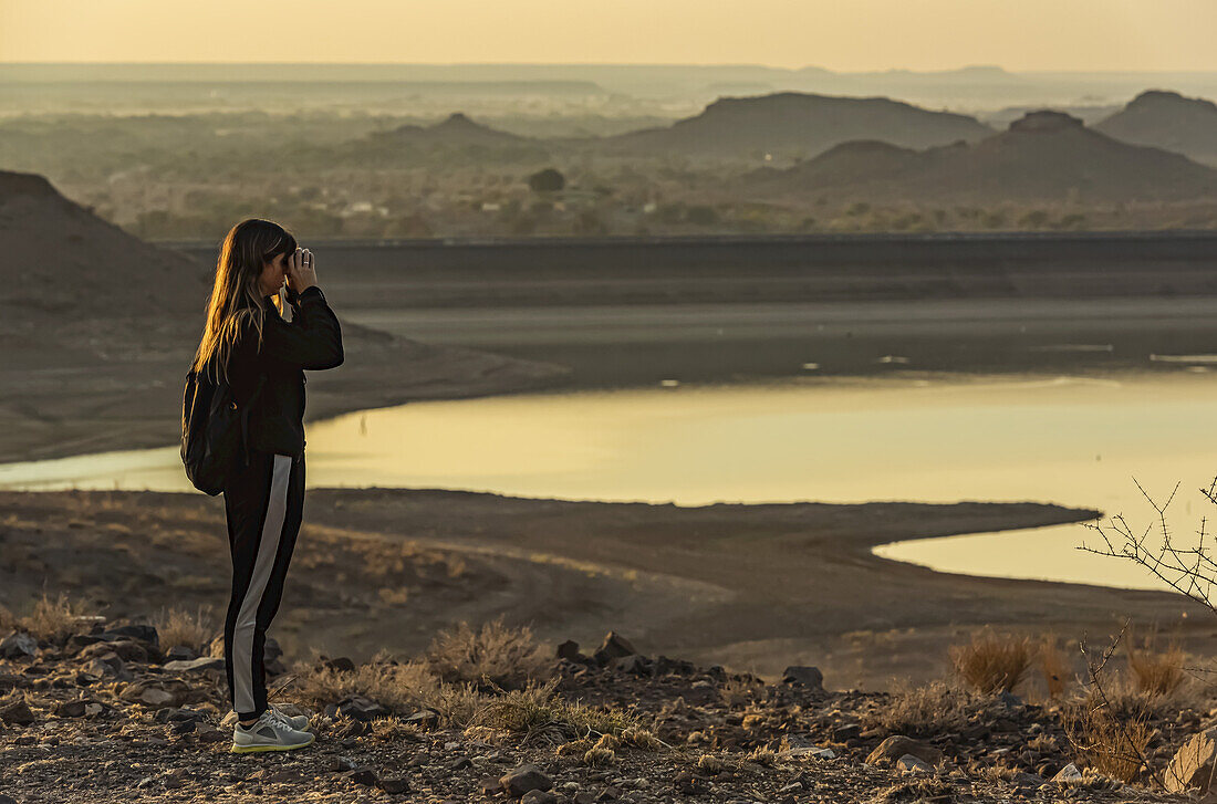 Weibliche Touristin schaut mit einem Fernglas bei Sonnenaufgang auf den Hardap-Damm; Hardap-Region, Namibia.