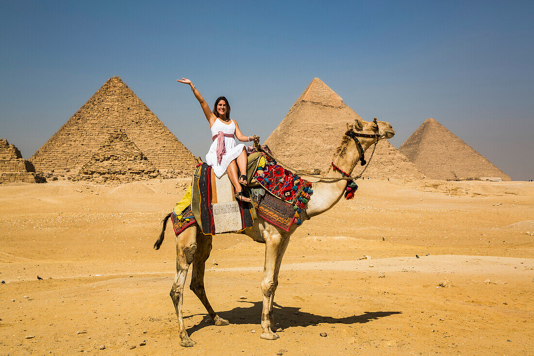 Weibliche Touristin winkt, während sie auf einem Kamel sitzt, Pyramidenkomplex von Gizeh, UNESCO-Welterbe; Gizeh, Ägypten.