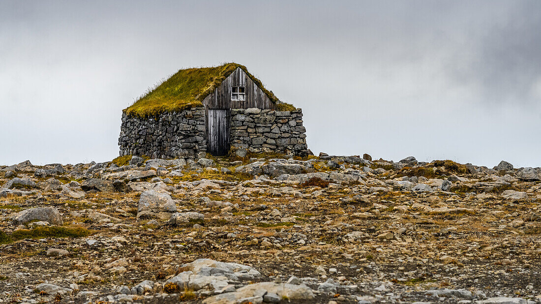 Kleines Gebäude aus Stein und Holz mit Grasdach am Horizont eines felsigen Geländes gegen einen bedeckten Himmel; Sudavik, Westfjorde, Island