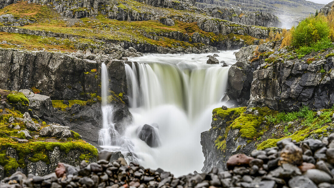 Ein Wasserfall über einer felsigen Landschaft in Herbstfarben; Djupivogur, östliche Region, Island.