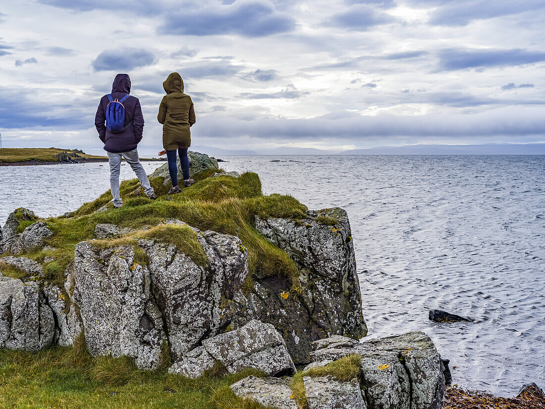 A man and woman tourist stand on a rocky ridge overlooking the ocean and coastline in Northern Iceland; Hunathing vestra, Northwestern Region, Iceland