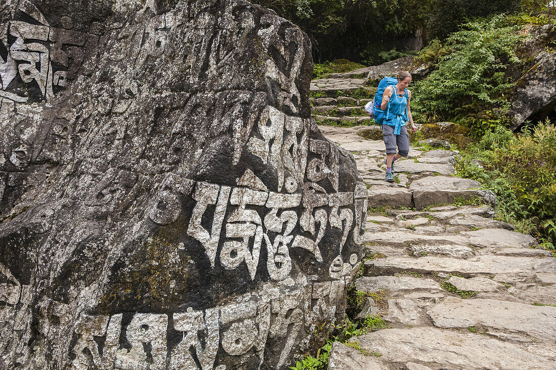 A woman wearing a backpack walks down a stone trail, pat a rock wall with script on it; Nepal