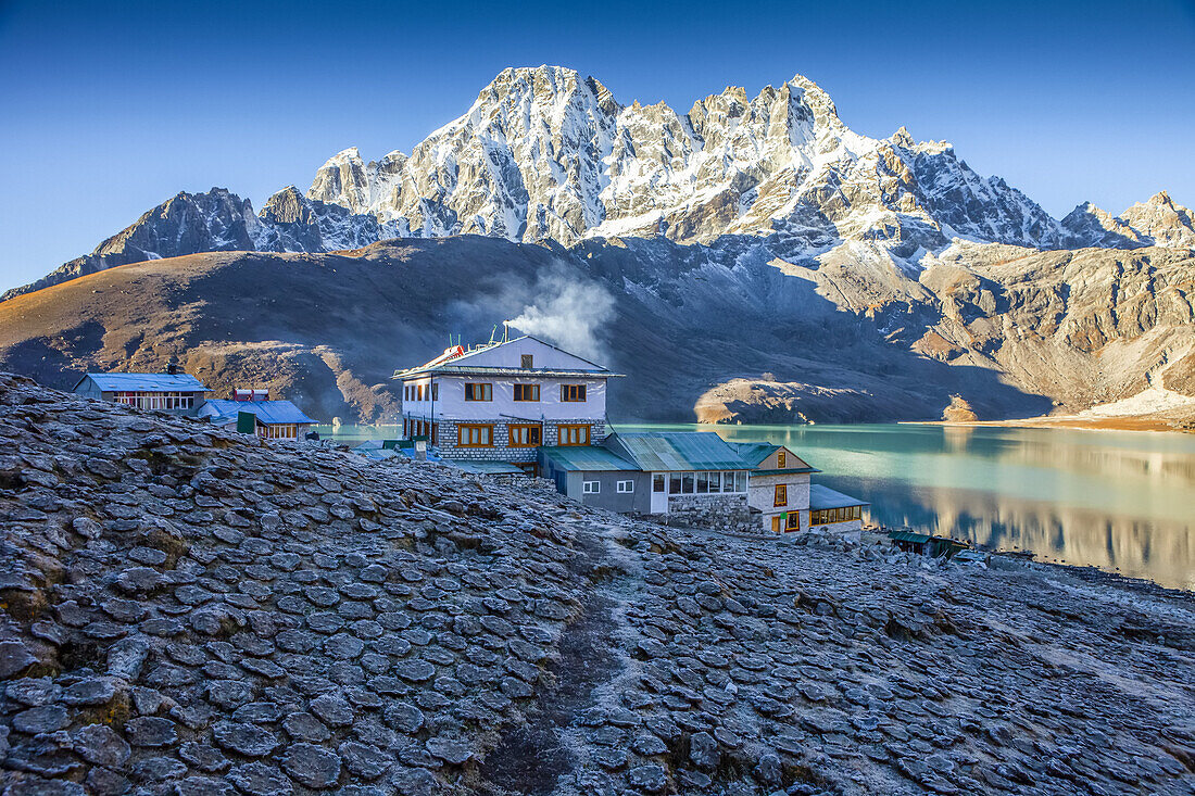Trail to the Village of Gokyo, surrounded by frosty yak dung patties set out to dry to be burned and used as heat for the teahouses, with the sacred turquoise colored Gokyo Lake and snow covered Himalayan peaks against a the blue sky on sunny autumn day; Sagarmatha National Park, Solokhumbu District, Nepal
