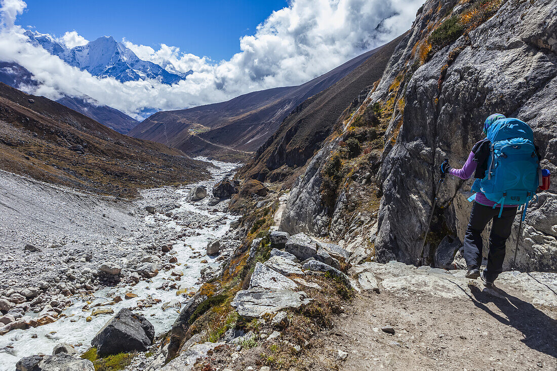 Backview of Caucasian woman trekker carrying blue backpack hiking down the Gokyo Trek trail, next to the rushing Dudh Kosi river towards a snow capped mountain in the distance on a sunny autumn day in Sagarmatha National Park; Solokhumbu District, Nepal