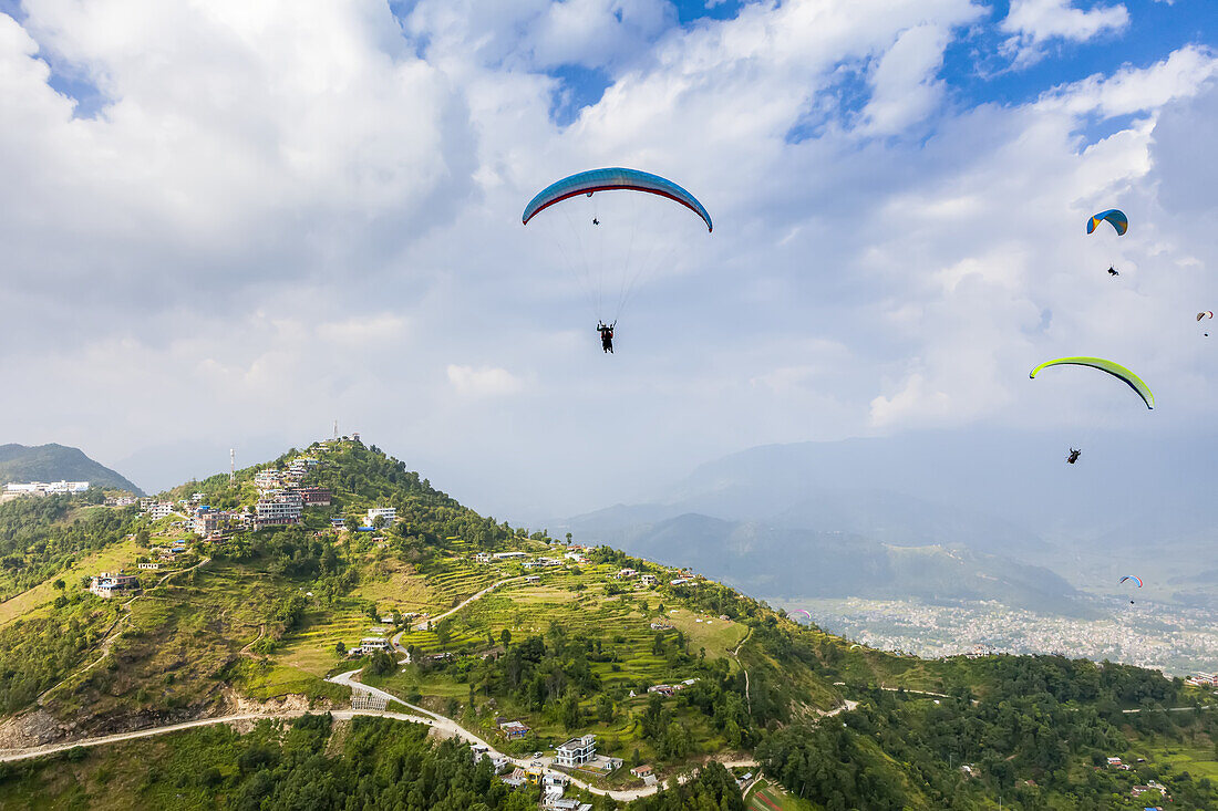 A group of colorful paragliders playing in the thermals above the terraces of Sarangkot with the city of Pokhara in the distance, on a sunny autumn day with a cloudy sky; ?Kaski District, Nepal