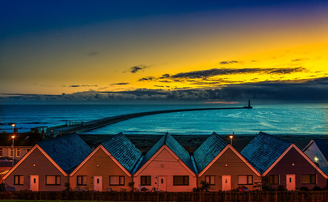 Beleuchtete Häuserreihe am Roker Beach in Sunderland mit einer Silhouette des Roker-Leuchtturms in der Ferne am Ende des Piers und des langen Wellenbrechers, der bei Sonnenuntergang aus dem Ufer herausragt; Sunderland, Tyne And Wear, England, Vereinigtes Königreich.