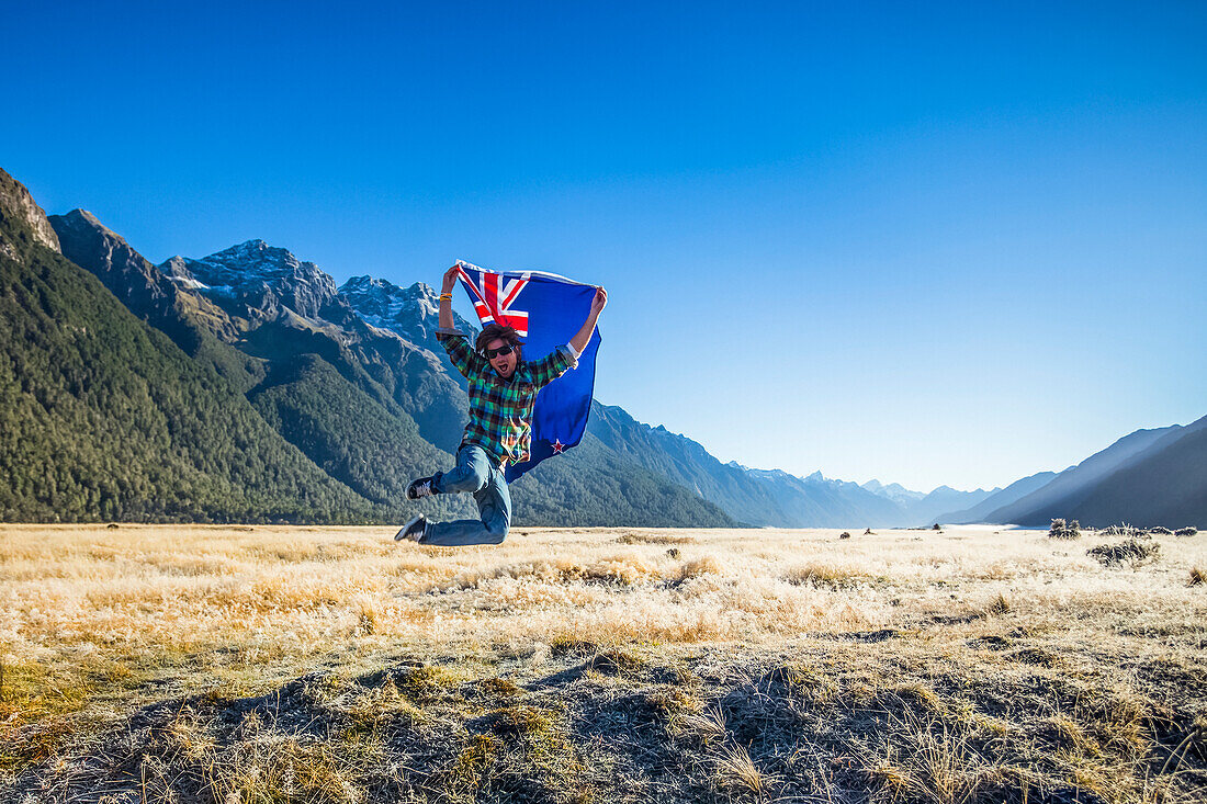 A man jumps in the air with the New Zealand flag in Fiordland National Park; Southland, New Zealand