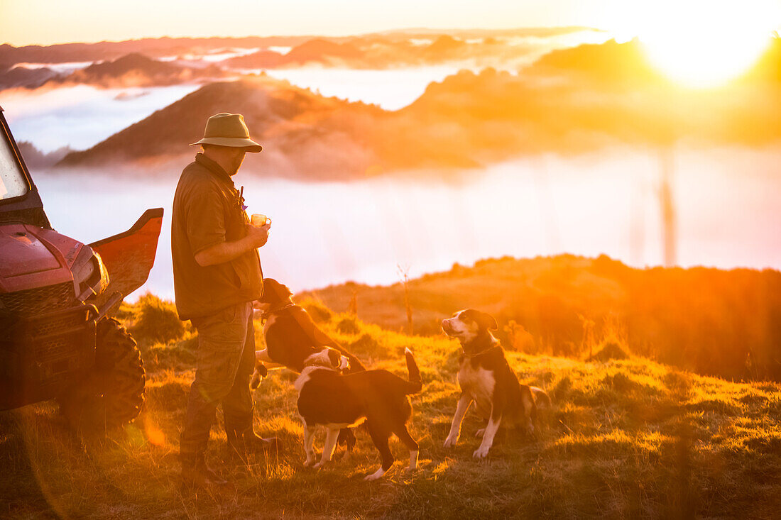 The Blue Duck lodge located in the Whanganui National Park is a working cattle farm with a focus on conservation. Travellers go to a scenic viewpoint to watch the sunrise over the rainforest. A man stands drinking coffee with dogs at his feet; Retaruke, Manawatu-Wanganui, New Zealand