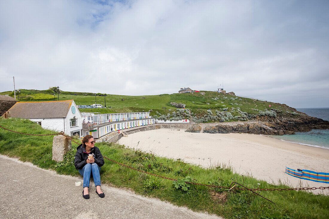 Eine junge Frau sitzt mit einer Kaffeetasse da und blickt auf den Strand und die Küste von St. Ives, einem kleinen Fischerdorf an der Südwestspitze Cornwalls, England. Bunte Strandhotels liegen an der Küste; Saint Ives, Cornwall, England
