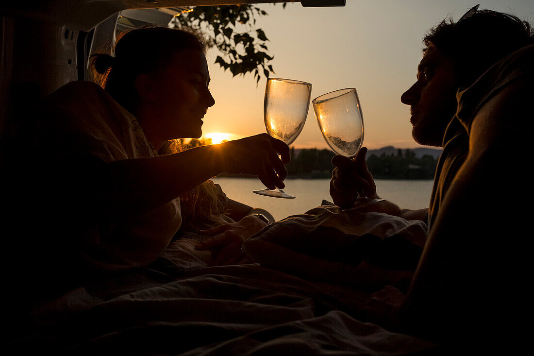 Ein junges Paar prostet sich mit Weingläsern auf dem Rücksitz eines Wohnmobils bei Sonnenuntergang zu. Eine Gruppe von Reisenden fährt an einem See in Bratislava vor, um dort zu übernachten; Bratislava, Region Bratislava, Slowakei