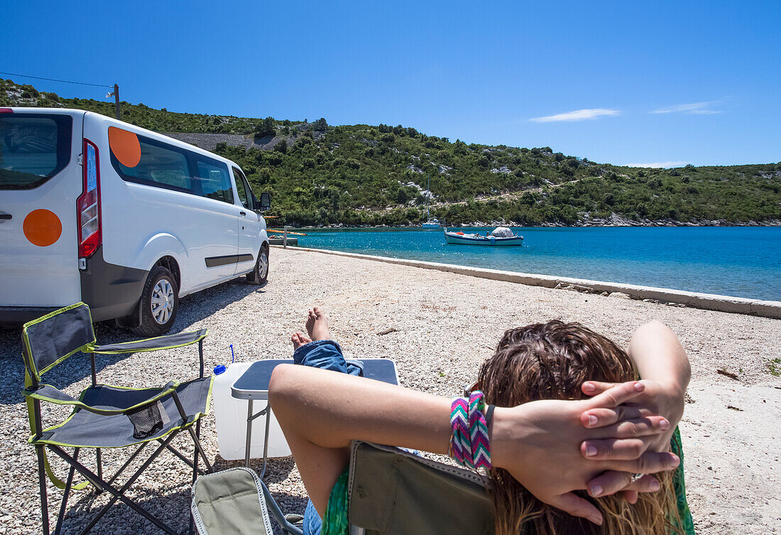 Frau sitzt in der Sonne und ruht sich außerhalb eines Wohnmobils aus, als eine Gruppe von Reisenden in Slano für einen Nachmittag am Strand anhält; Slano, Gespanschaft Dubrovnik-Neretva (Dubrova?ko-neretvanska ?upanija), Kroatien
