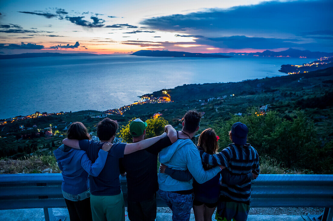 Blick von hinten auf Reisende, die zusammenstehen und die Arme umeinander legen, während sie an der Straße anhalten, um den Sonnenuntergang zu beobachten. Die atemberaubende Aussicht auf die hoch gelegenen Klippen entlang der kroatischen Küste; Podgora, Gespanschaft Dubrovnik-Neretva (Splitsko-dalmatinska zupanija), Kroatien