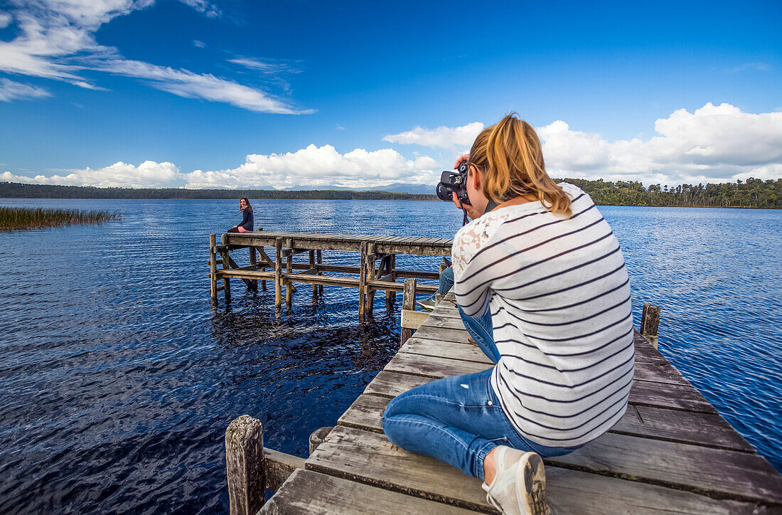 Blick von hinten auf eine Frau, die eine andere Frau auf einem Holzsteg fotografiert, während sie den Lake Mahinapua erkundet; Westküste, Neuseeland.