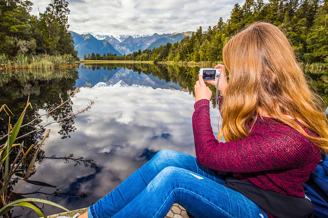 Tourist erkundet die Lake Matheson Mirror Lakes. Der wolkenverhangene Himmel verwässert die Spiegelung an diesem Tag etwas; South Westland, Neuseeland