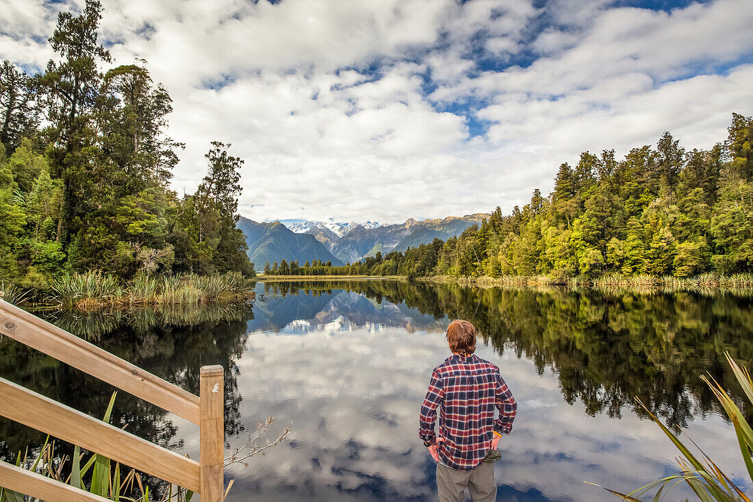Tourist erkundet die Lake Matheson Mirror Lakes. Der bewölkte Himmel verwässert die Spiegelung an diesem Tag etwas; South Westland, Neuseeland