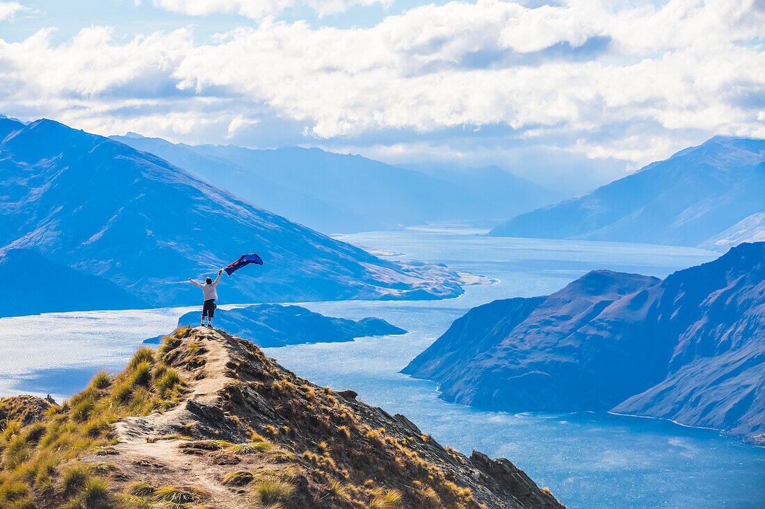 The strenuous yet highly rewarding hike to Roys Peak in Wanaka. The hike is difficult but the views are spectacular. A traveler celebrates at the summit by waving a New Zealand flag; Wanaka, Otago, New Zealand