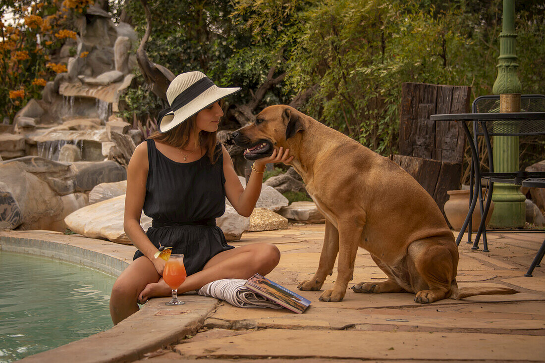 Frau mit Sonnenhut sitzt an einem Swimmingpool und streichelt einen Hund (Canis lupus familiaris) auf der Gabus Game Ranch; Otavi, Otjozondjupa, Namibia.
