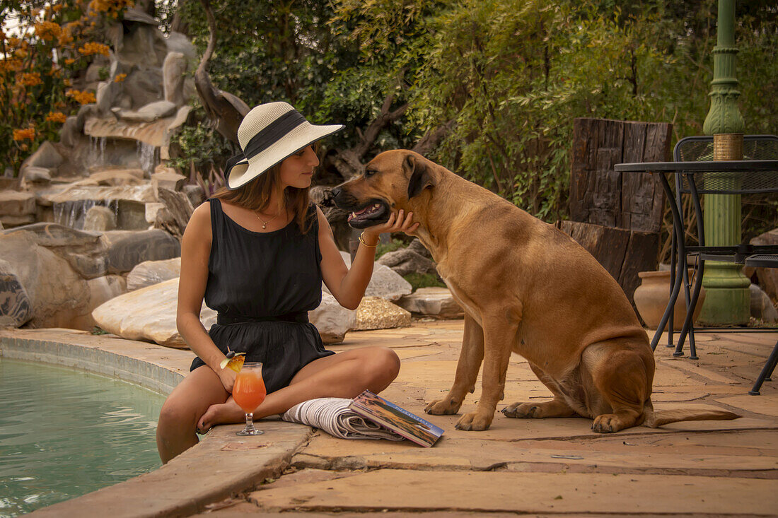 Woman wearing sunhat sitting by a swimming pool and petting a dog (Canis lupus familiaris) at the Gabus Game Ranch; Otavi, Otjozondjupa, Namibia