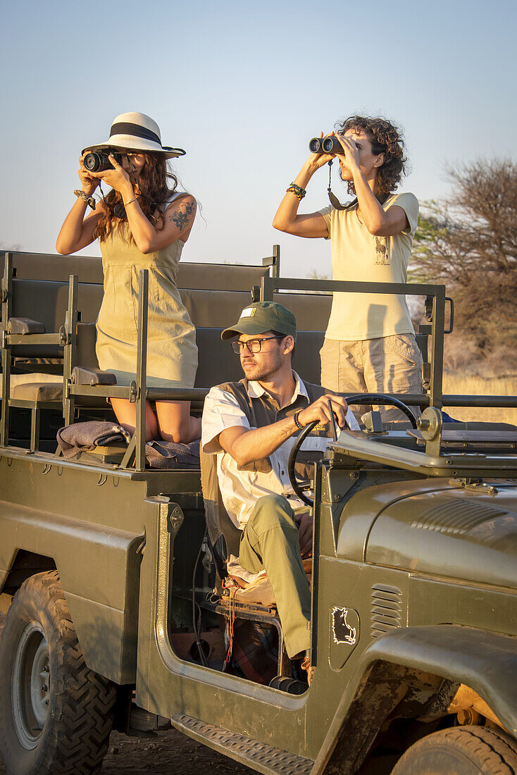 Nahaufnahme von Frauen in einem Jeep, eine mit einem Fernglas und eine mit einer Kamera, die in die Savanne blicken, während der Fahrer hinter dem Lenkrad sitzt, während einer Safari auf der Gabus Game Ranch; Otavi, Otjozondjupa, Namibia.