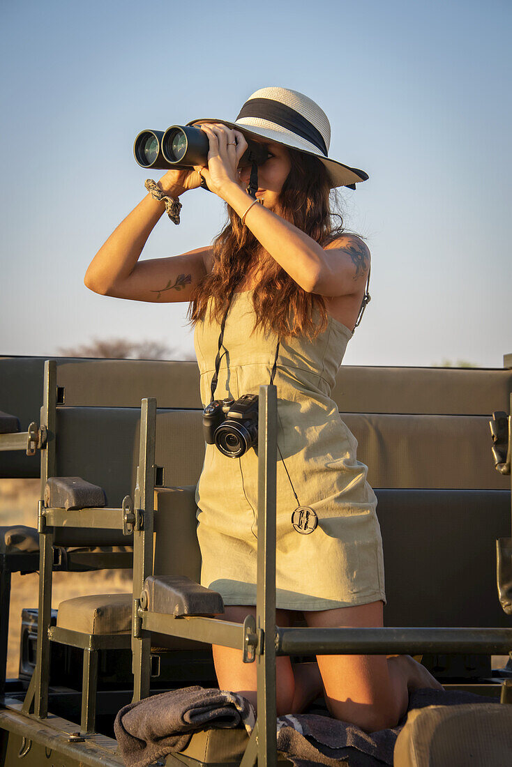 Nahaufnahme einer Frau, die in einem Jeep auf Safari kniet, einen Strohhut trägt, eine Kamera um den Hals hat und mit einem Fernglas auf die Savanne der Gabus Game Ranch bei Sonnenuntergang schaut; Otavi, Otjozondjupa, Namibia.