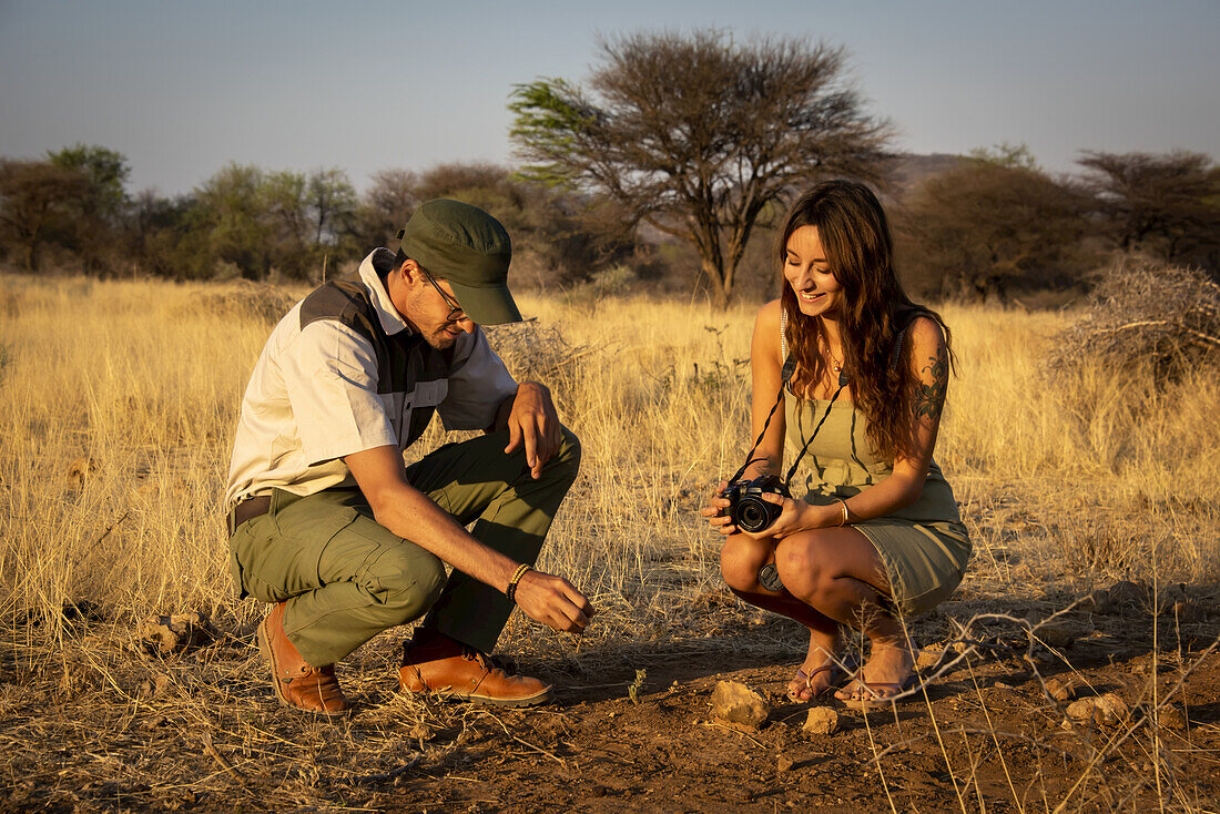 Safari-Führer mit einer Reisenden, die eine Kamera in der Hand hält, kauert auf dem Boden und unterhält sich in der Savanne auf der Gabus Game Ranch bei Sonnenuntergang; Otavi, Otjozondjupa, Namibia.