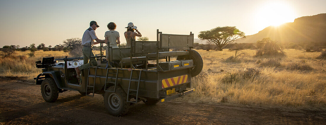 Blick von hinten auf einen Safari-Führer und eine Reisende, die in einem geparkten Jeep stehen, sich unterhalten und mit einem Fernglas auf die Savanne auf der Gabus Game Ranch bei Sonnenaufgang schauen; Otavi, Otjozondjupa, Namibia.