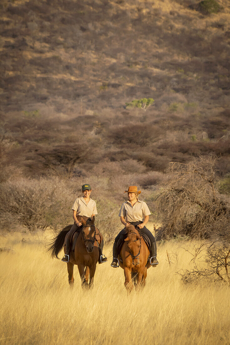 Two women riding horses (Equus ferus caballus) traveling through the grass near a hill at the Gabus Game Ranch at sunset; Otavi, Otjozondjupa, Namibia