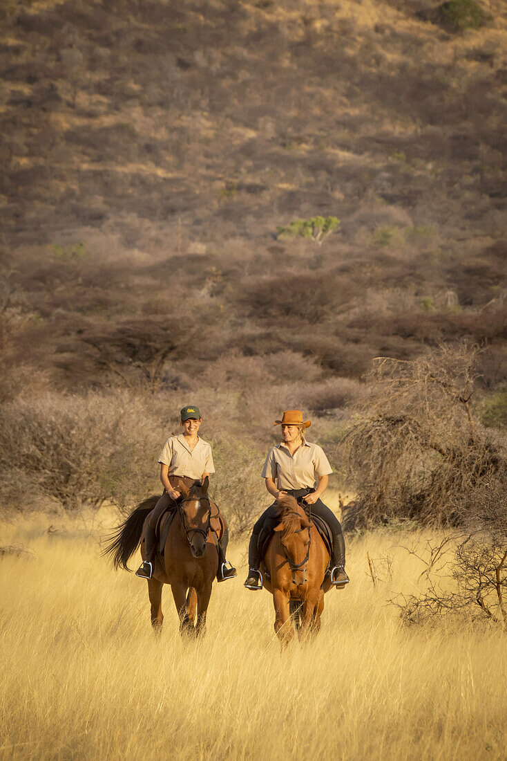 Zwei Frauen reiten auf Pferden (Equus ferus caballus), die durch das Gras in der Nähe eines Hügels auf der Gabus Game Ranch bei Sonnenuntergang reiten; Otavi, Otjozondjupa, Namibia