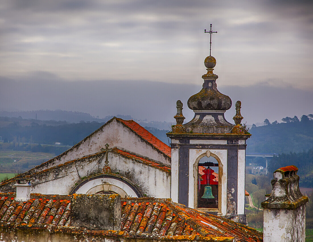 Old bell tower and clay tiled rooftops overlooking the countryside and the medieval town of Obidos with a hazy, overcast sky; Obidos, Estremadura, Oeste Region, Portugal