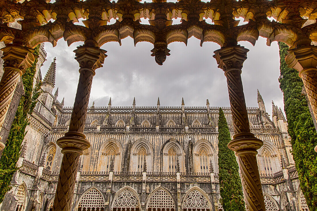Looking out through the ornate columns of the cloister up at the grey clouds into the inner courtyard towards the chapel at the medieval Monastery of Batalha, a masterpiece of Gothic architecture; Batalha, District of Leiria, Centro Region, Portugal