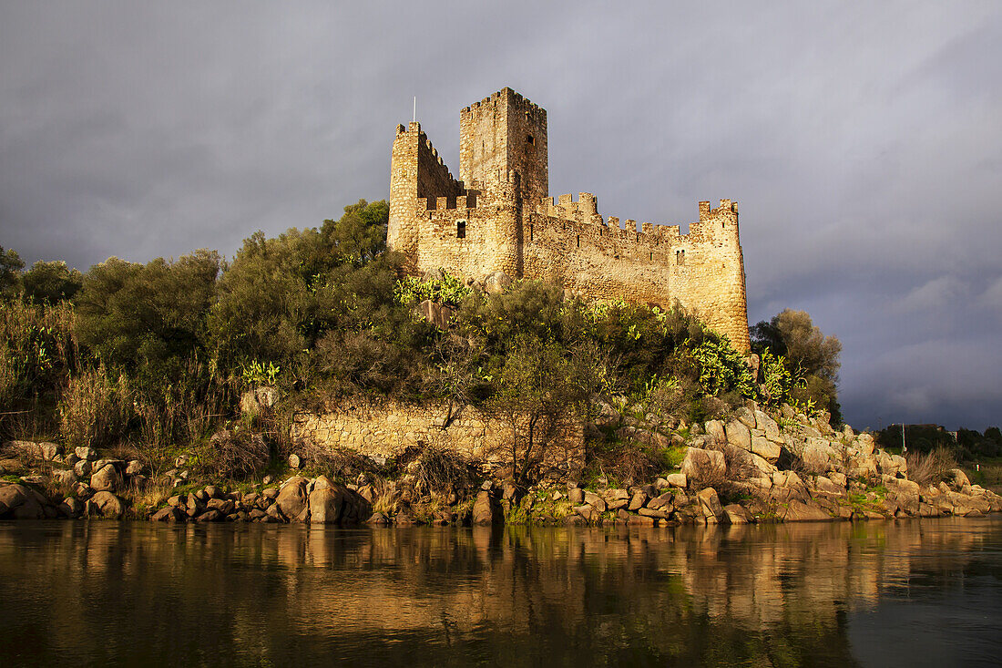 Warm sunlight reflecting on the historical Castlo de Almourol on the islet of Almourol in the River Tagus with a grey sky; Vila Nova da Barquinha, Praia do Ribatejo, Centro Region, Portugal