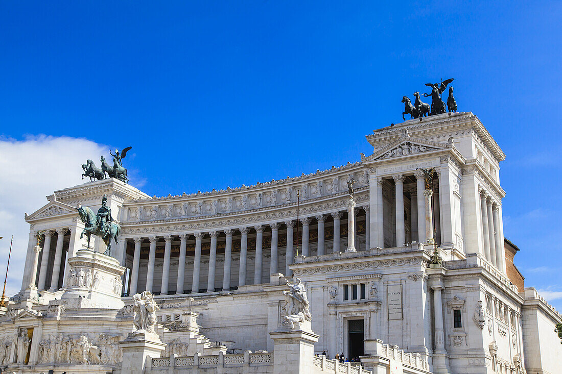 The iconic Victor Emmanuel II National Monument in white marble against a blue sky with a bronze, equestrian sculpture of Victor Emmanuel II in the center and two statues of goddess Victoria on quadrigas on either side of the rooftop representing unity and freedom; Rome, Lazio, Italy