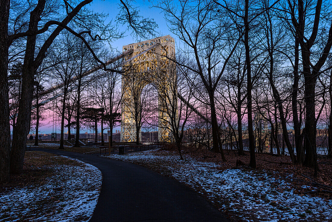 George Washington Bridge (specially lit for Martin Luther King Jr. Day) seen through the silhouetted bare trees along the pathway at the Fort Lee Historic Park at dusk; Fort Lee, New Jersey, United States of America