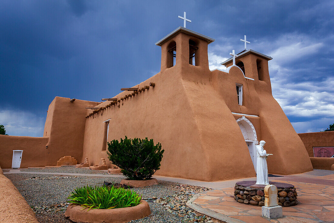 Seitenansicht der historischen Kirche San Francisco de Asis mit einer Statue von San Francisco de Asis im begrünten Innenhof auf dem Hauptplatz von Ranchos de Taos; Taos, New Mexico, Vereinigte Staaten von Amerika.