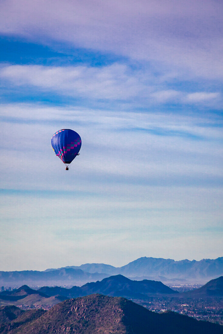 Blauer und violett gemusterter Heißluftballon in der Mitte des Fluges, der die silhouettierten Berge vor einem bewölkten, blauen Himmel überquert; Phoenix, Arizona, Vereinigte Staaten von Amerika.