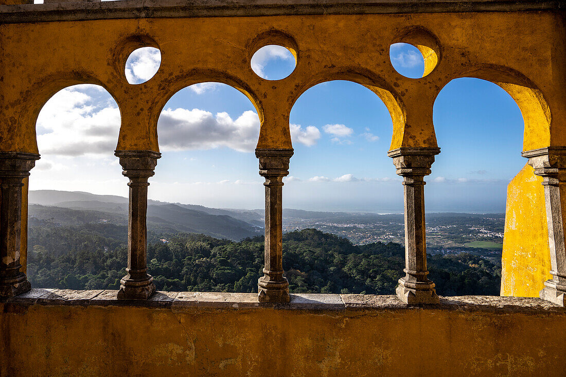 Blick durch die farbenfrohen Bögen und Säulen auf der Königinnenterrasse mit Blick auf die bewaldeten Anlagen des Pena-Parks und die Sintra-Berge, die das auf dem Hügel gelegene Schloss Palacio Da Pena umgeben; Sintra, Bezirk Lissabon, Portugal.