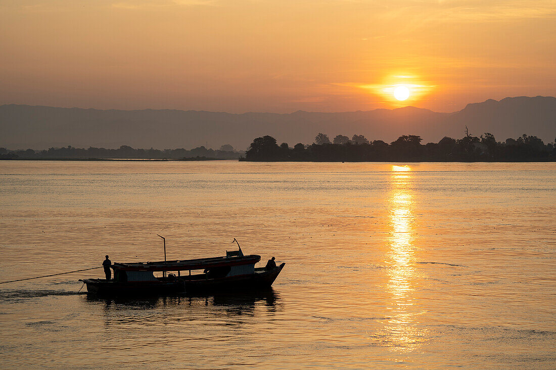 Silhouette eines Kajütboots, das bei Sonnenuntergang den Fluss hinauffährt; Ländlicher Dschungel, Mandalay, Myanmar (Burma)