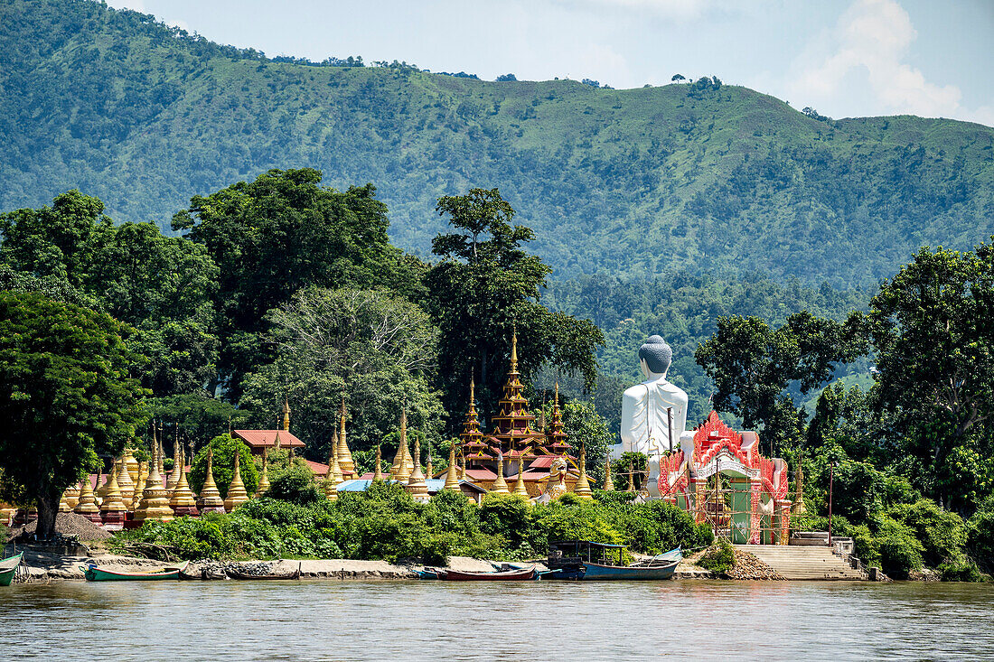 Blick von hinten auf einen Riesenbuddha neben Pagoden und goldenen Stupas auf der Insel Shwe Paw und Booten, die entlang des Ayeyarwady (Irrawaddy) Flusses vertäut sind; Shwegu, Kachin, Myanmar (Burma)