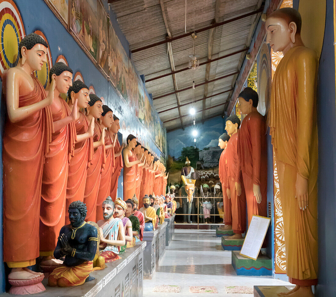 Flur gesäumt von Statuen stehender Buddhas und Skulpturen heiliger Männer, die vor ihnen sitzen, im buddhistischen Kloster Galagoda Shailatharama Viharaya; Balapitiya, Galle District, Sri Lanka.